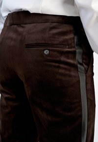 Nærbillede af mørkebrune fløjlsshorts med en knaplukket baglomme og sort satinstripe på siden, båret med en hvid skjorte.
