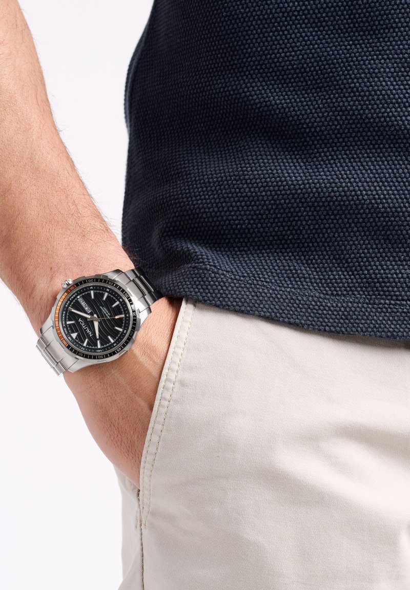 Reloj de acero inoxidable con un dial texturizado negro, acentos naranjas y pulsera plateada. Lleva puesto en una muñeca; camisa gris parcialmente visible.
