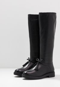 Bottes noires en cuir montantes jusqu'aux genoux, avec une texture lisse, des pampilles décoratives et une semelle robuste. Elles présentent un bout rond et une bordure cousue.