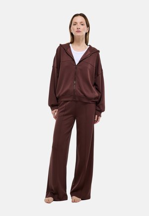 Femme debout pieds nus portant un pantalon large marron et un sweat à capuche zippé assorti avec un t-shirt blanc en dessous, fond clair uni.