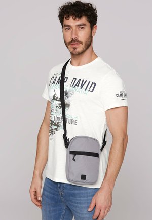 Gray crossbody bag with zip closure, a front pocket with black accent, worn over a white printed t-shirt, model in blue jeans.