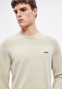 Világos bézs pulóver texturált anyagból, vízszintes bordázott részletekkel a vállakon. Kisméretű fekete "BOSS" logóval.