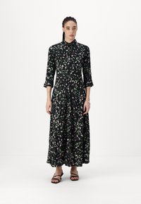 Robe noire longue avec un motif de feuilles vertes et roses, devant boutonné, col traditionnel et manches trois quarts. Portée avec des sandales noires à lanières.
