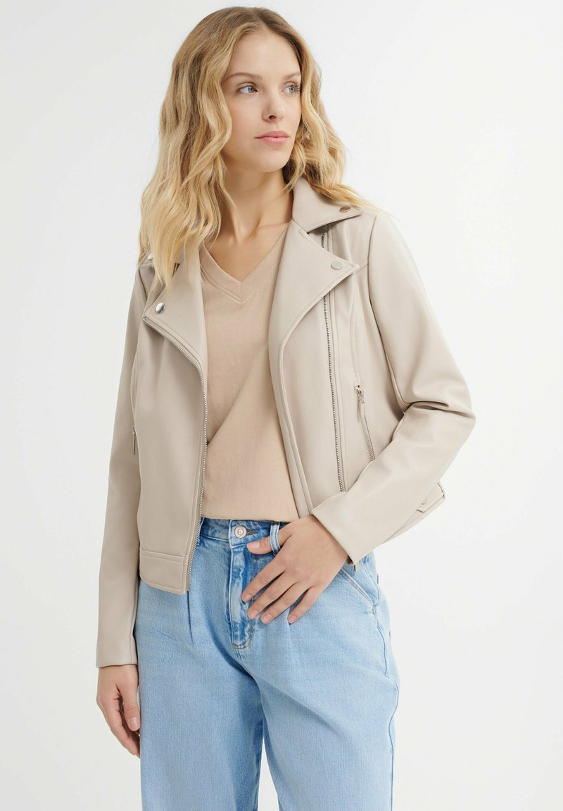 Giacca da motociclista in ecopelle beige con tasche con zip, hardware argentato e silhouette aderente, abbinata a un top beige e jeans azzurri chiari.