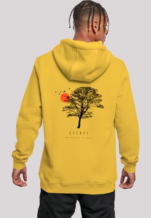 Person, der einen senfgelben Hoodie mit schwarzem Baum, orangefarbener Sonne und fliegenden Vögeln auf dem Rücken trägt, steht vor einem schlichten Hintergrund.