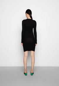 Abito a coste nero con maniche lunghe, caratterizzato da una silhouette aderente e una lunghezza a metà coscia. Modello che indossa mule con tacco blocco verde.