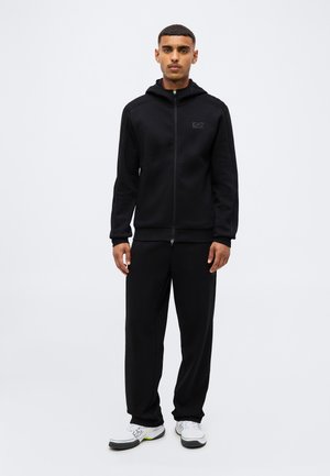 Jeune homme portant une veste à capuche noire zippée et un pantalon noir avec des baskets blanches, debout devant un fond clair uni.
