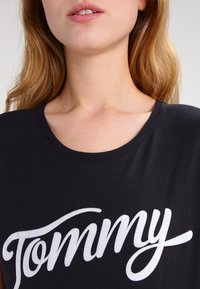 Camiseta de algodón negra con escote redondeado, que presenta un logo en cursiva blanca "Tommy" en el pecho. Textura suave con mangas cortas.