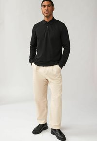 Zwarte langwerpige poloshirt met een kraag, gecombineerd met lichte beige geplooide broeken en zwarte instappers, met een gladde textuur en een minimalistisch ontwerp.