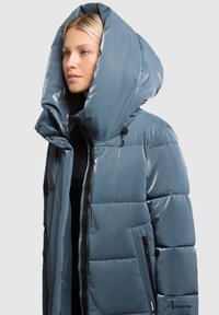 Pehmustettu takki kiiltävästä teräksen sinisestä materiaalista, jossa on ylisuuri huppu. Varustettu vetoketjullisilla sivutaskuilla ja etuosan tarrakiinnityksellä.