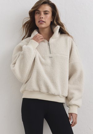 Jeune femme aux cheveux ondulés porte un pull sherpa polaire de couleur crème avec une fermeture éclair à l'avant et des poignets côtelés, assorti à des leggings noirs.