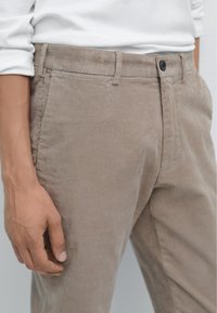 Pantaloni in corduroy beige con texture liscia, caratterizzati da una chiusura con bottone e una vestibilità sartoriale. Passanti per cintura visibili e tasche laterali.