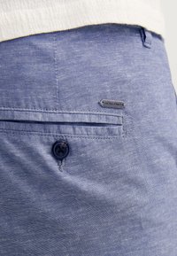 Pantalones cortos de tela azul claro con una textura sutil, que cuentan con un cierre de botón y una pequeña etiqueta de marca cerca del bolsillo trasero.