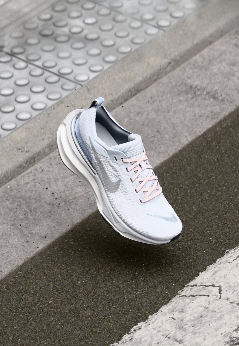 Chaussure de sport blanche avec le logo Nike gris et des lacets rose pâle sur un trottoir urbain près du bord du trottoir et du pavage tactile.