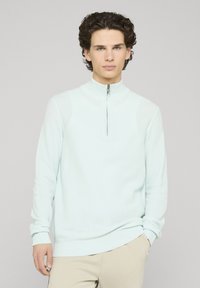 Pull bleu clair zippé avec un col haut, des poignets et un ourlet côtelés, fabriqué en tissu tricoté texturé. Coupe décontractée avec poches latérales.