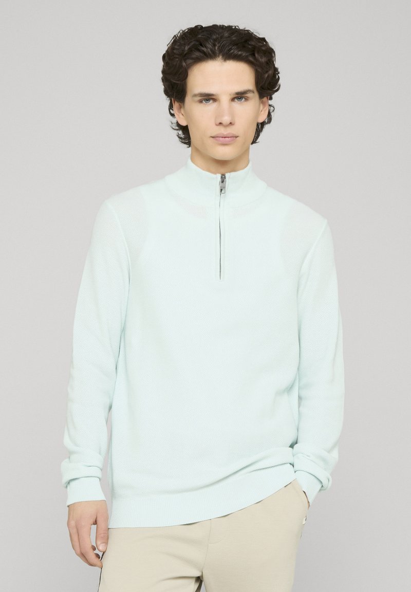 Pull bleu clair zippé avec un col haut, des poignets et un ourlet côtelés, fabriqué en tissu tricoté texturé. Coupe décontractée avec poches latérales.