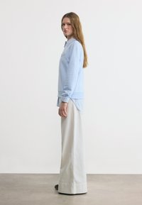 Νεαρή γυναίκα με ανοιχτό μπλε φούτερ πάνω από ριγέ πουκάμισο, φαρδιά λευκά παντελόνια, σε πλάγια στάση δίπλα σε απλό λευκό τοίχο.