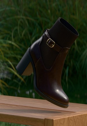 Stivaletto alla caviglia in pelle marrone scuro con tacco spesso, polsino in tessuto rigato e cinturino con fibbia dorata, sospeso sopra una superficie in legno all'aperto.