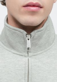 Maglione grigio con zip e collo alto, caratterizzato da una texture liscia e una zip metallica al collo. Vista ravvicinata del tessuto.