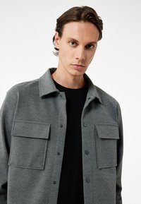 Grau button-up Hemdjacke aus strukturiertem Stoff, mit zwei Brusttaschen, einem Kragen und schwarzen Druckknöpfen. Über einem schwarzen Hemd getragen.