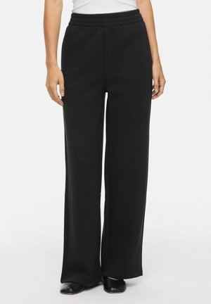 Personne portant un pantalon noir à jambes larges avec une taille élastique et des chaussures noires, visible de la taille aux pieds sur un fond uni.