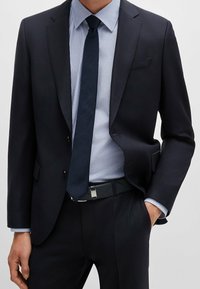 Námořnicky modrý oblek z texturované látky, kombinovaný s světle modrou pruhovanou košilí a tmavou kravatou. Součástí je černý pásek se stříbrnými detaily.