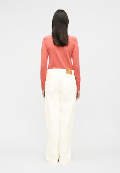 Pikkade tumejate juustega naine, kes kannab korallivärvi pika varrukaga särki ja kõrge vöökohaga valgeid laia säärega pükse, seisab selja poole.