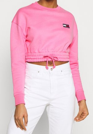 Rosa cropped sweatshirt med snor i kanten, ribbede manchetter og et lille logo patch. Parret med hvide denim bukser. Blød stoftekstur.