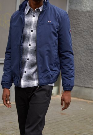 Homme portant une veste bleu marine par-dessus une chemise à carreaux noire et blanche boutonnée et un pantalon noir, marchant sur un trottoir urbain.