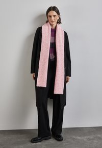 Écharpe longue rose côtelée en matériau doux, portée sur un pull en tricot violet à motifs et un manteau noir, associée à un pantalon noir et des chaussures noires.
