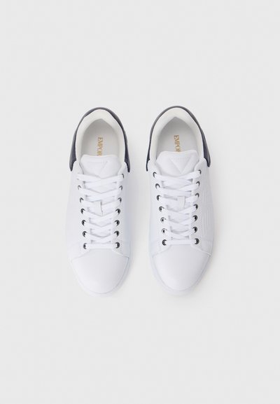 Et par hvite sneakers med svarte hæltabber, hvite lisser og gull "EMPOR" merking på innersålene, sett ovenfra på en hvit bakgrunn.