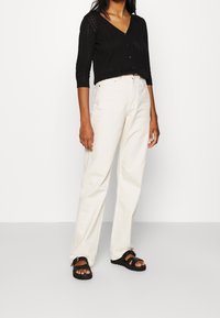 Femme portant un cardigan noir à motif avec col en V, un pantalon droit taille haute couleur ivoire, et des sandales noires à bout ouvert avec des boucles.