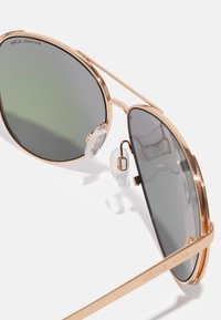 Ochelari de soare tip aviator cu rame din metal roz aurit, lentile gri-verzui cu degradé, pernițe nazale ajustabile și gravură vizibilă a mărcii pe brăcat.