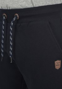 Navy blue sweatpants in soft fabric feature a drawstring waistband with contrasting white patterns and a brown leather logo patch on the side.