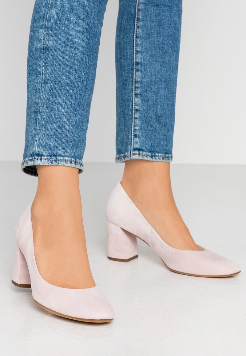 Escarpins à talons hauts en daim rose clair avec un bout arrondi, dotés d'un talon bloc et d'une texture lisse, associés à un jean bleu longueur cheville.