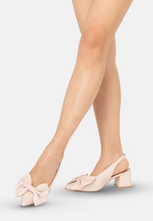 Beine, die hellrosa Slingback-Schuhe mit niedrigem Blockabsatz und großen dekorativen Schleifen vorne tragen, vor einfachem Hintergrund.