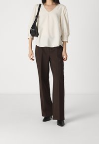 Lichtbeige V-hals blouse met poffende mouwen en kanten afwerking, gecombineerd met donkerbruine hoge taille broek en zwarte haklaarsjes.