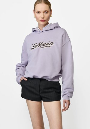 Fioletowa bluza z kapturem z haftem "LaMania" w ciemnym kolorze, luźny krój, opadające ramiona, zestawiona z czarnymi szortami w prążki. Gładka faktura.