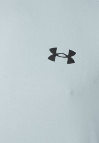 Ljusblå idrottströja i slätt tyg. Har en framträdande svart Under Armour-logotyp på bröstet. Klassisk rundhalsad design.