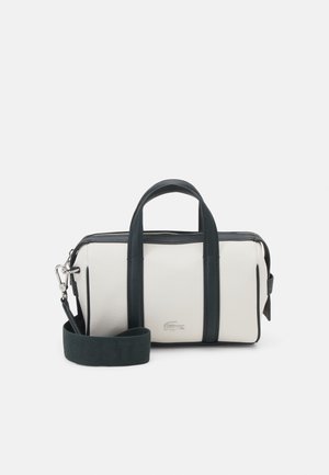 Petit sac rectangulaire blanc avec des poignées noires, une bandoulière noire ajustable et une fermeture éclair argentée, présentant un petit logo crocodile brodé.