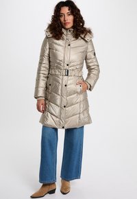 Vrouw in een beige gebeltste pufferjas met een bontomzoomde capuchon, blauwe wijde jeans en tan enkellaarsjes, staand tegen een witte achtergrond.