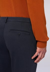 Närbild av en person som bär marinblå byxor med en knappad bakficka och en bränd orange långärmad tröja.