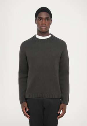Maglione girocollo grigio scuro a maglia con polsini e orlo a coste, caratterizzato da un motivo testurizzato. Indossato sopra una camicia bianca.