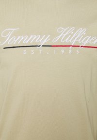 Lyhythihainen, vaaleanbeige puuvillapaita, jossa on brodeerattu "Tommy Hilfiger" -logo valkoisena punaisilla ja mustilla yksityiskohdilla, perustettu 1985.