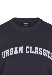 Tumši zils kokvilnas T-krekls ar noapaļotu apkakli un lielu balto uzdrukāto tekstu "URBAN CLASSICS" krūšu daļā.