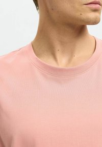 Ljusrosa t-shirt med kort ärm och rund halsringning. Tillverkad av slät, stretchig tyg. Synlig söm runt halsringningen och axlarna.