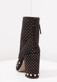 Musta mokkanahkainen nilkkabotti, jossa on vetoketjusulku, koristeena kultasävytteiset niitit ja tukeva korko, pyöreä varvas ja sileä sisäosa.