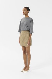Top gris à manches courtes avec un petit logo, associé à une jupe à carreaux jaunes et marron avec un bouton sur le côté. Le modèle porte des talons crème.
