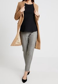 Cappotto lungo beige, top nero testurizzato, pantaloni grigi a quadri e ballerine nere. Fodera del cappotto liscia e visibile. Design aderente che enfatizza la silhouette.