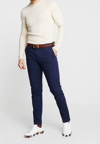 Granatowe spodnie o wąskim kroju, zestawione z kremowym, prążkowanym swetrem. Pasek wykonany z brązowej skóry. Białe trampki z wzorzystymi akcentami.